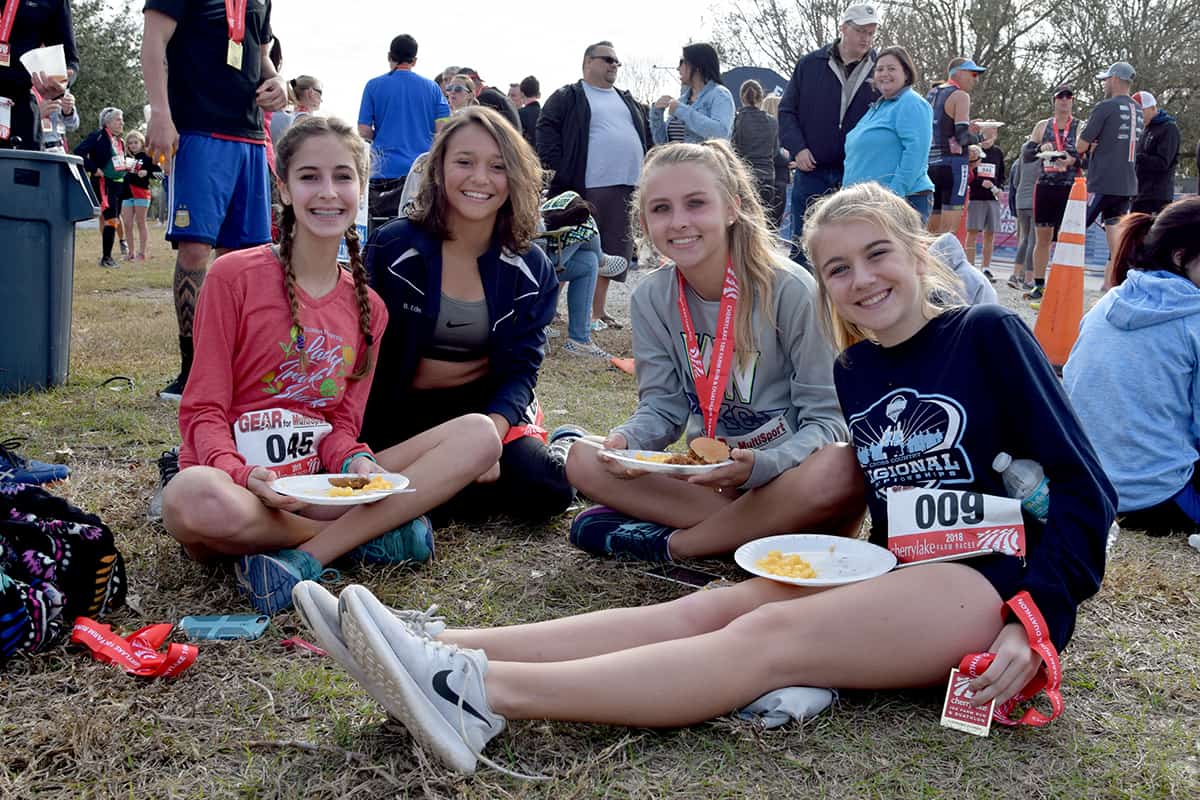 Four teenage girls smile sitting on the grass eating food after a race.