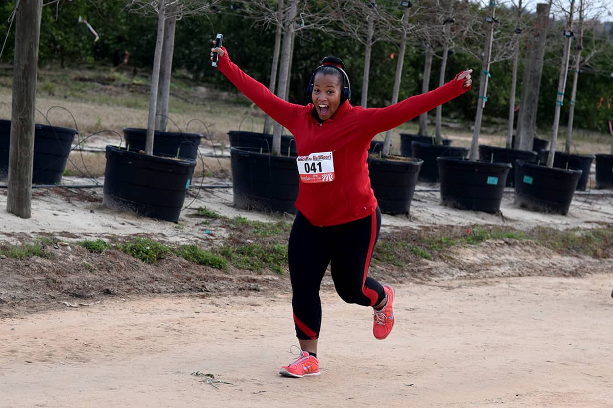 A woman holds her arms in a Y shape of excitement as she runs a race on an off-road path beside rows of containerized trees.