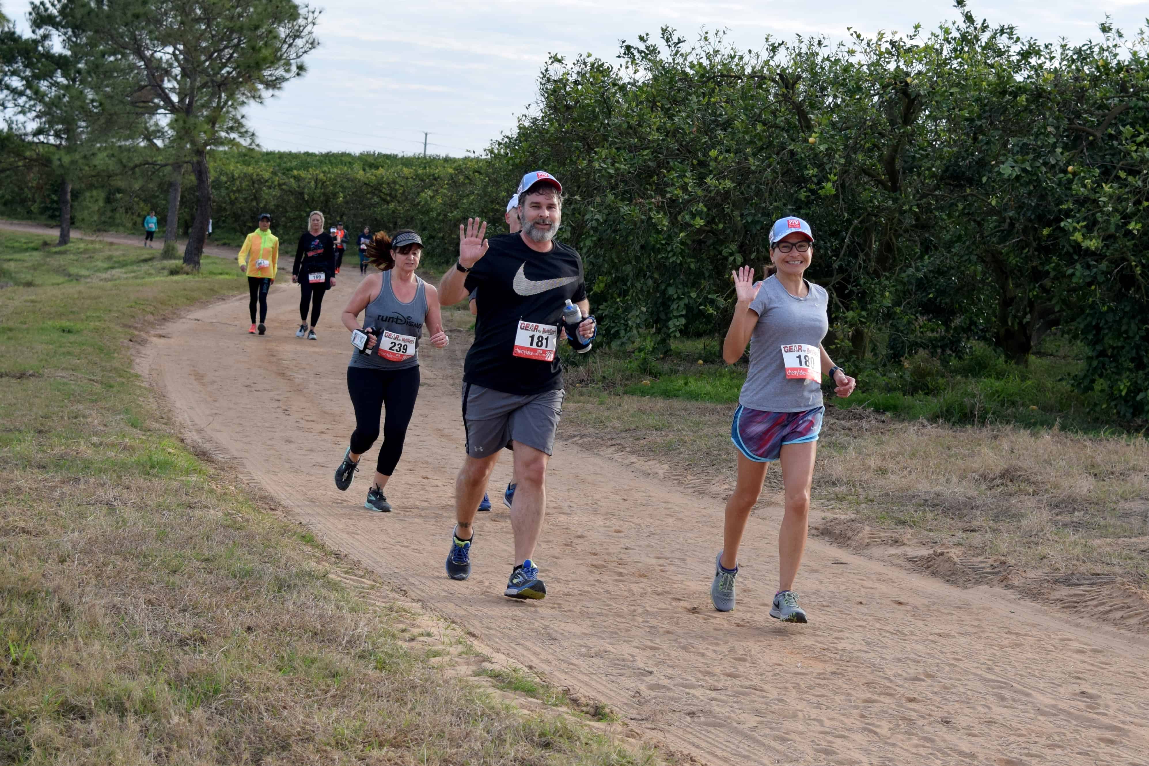 A couple running alongside each other wave to the camera as they run on an off-road trail surrounded by citrus trees.