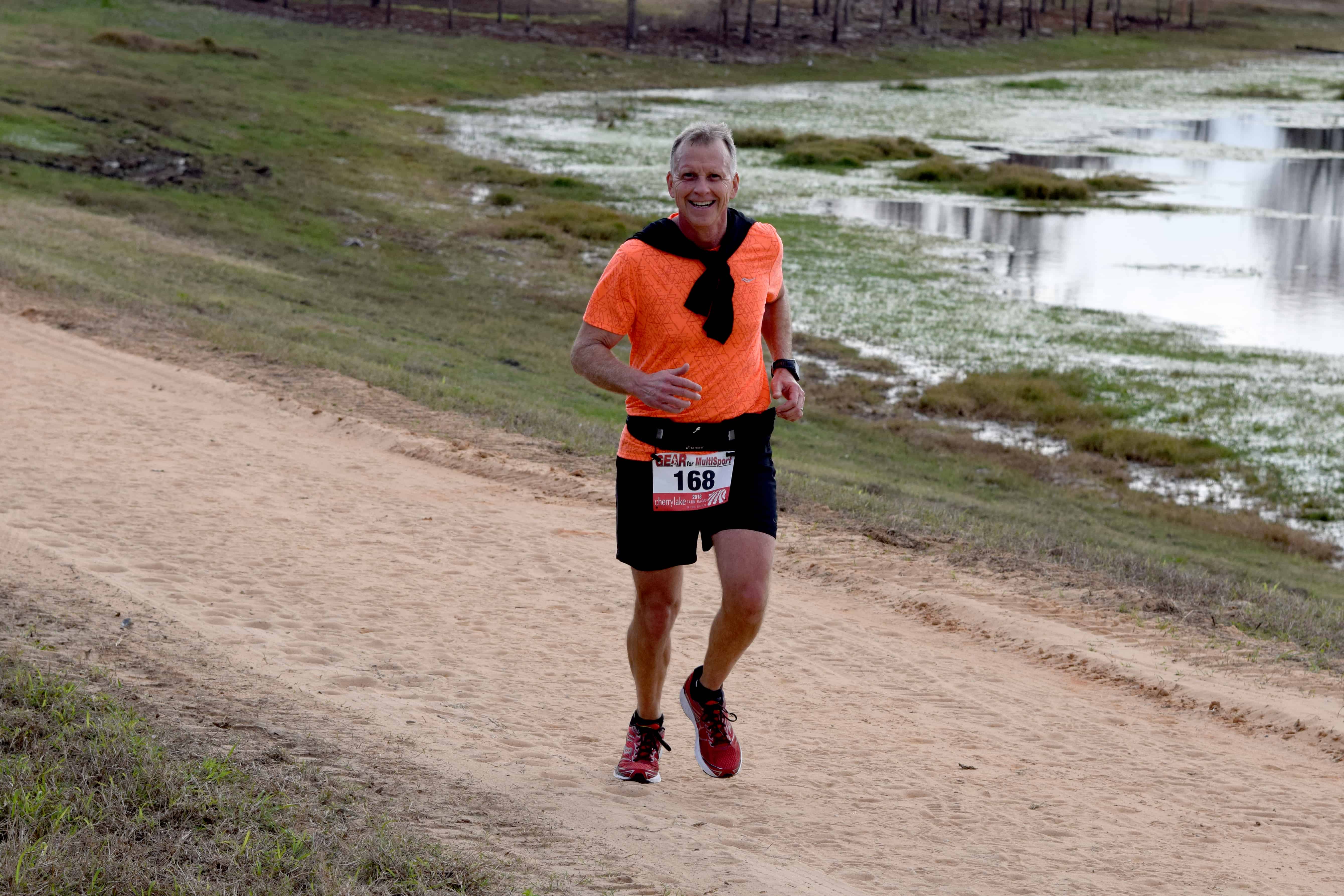 A man in an orange shirt smiles as he runs on an off-road path by a lake.