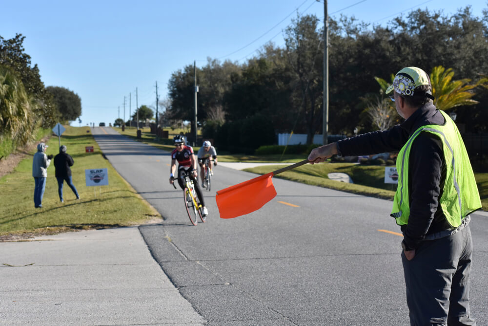 A man with a yellow safety vest directs the cyclists left using an orange flag.