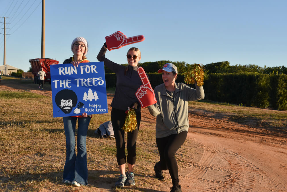 Three women encouraging runners, one holding a Bob Ross poster painted “Run for the trees, happy little trees” and the other two waving orange pointed fingers and golden pom poms.